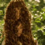 E, close-up image of a woman's hair, with a few strands lifted to reveal a delicate, ornate, antique-style hair oil bottle in the background, surrounded by lush greenery