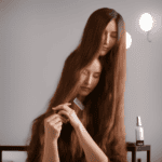 E, minimalist background with a beautiful, long-haired woman sitting in front of a vanity, surrounded by various hair oils and a comb, gently applying oil to the ends of her hair